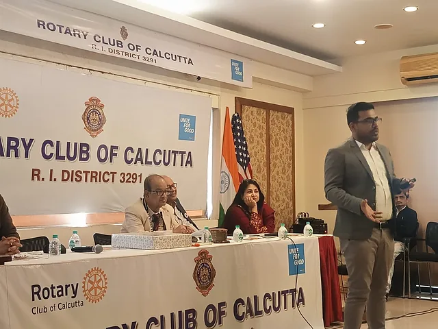 Presenter standing beside a speakers' table under a Rotary Club of Calcutta banner