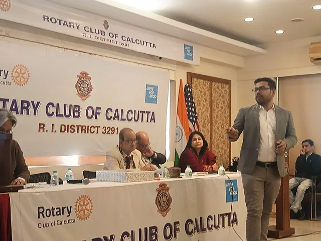 Presenter addressing the audience beside a speakers' table with flags and a banner
