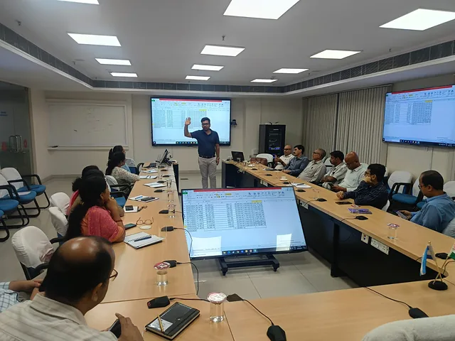Presenter raising a hand toward wall-mounted screens while participants listen around a boardroom table